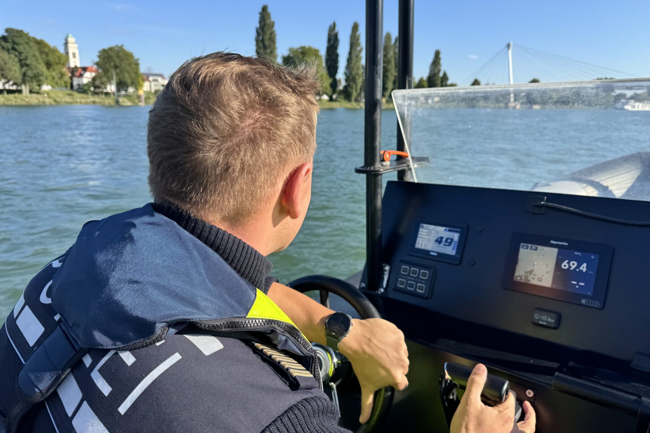 Person steuert Boot, Blick auf Wasser und Ufer mit Bäumen.