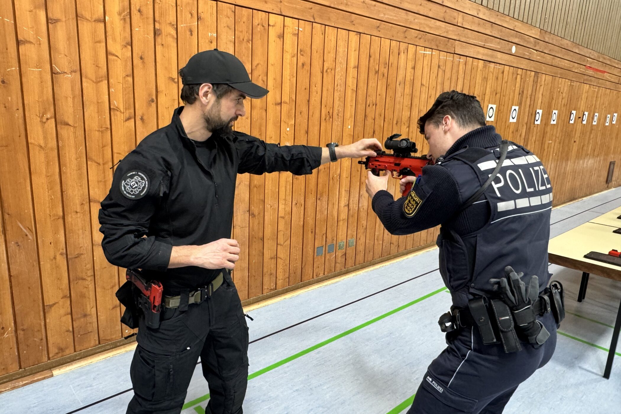 Zwei Polizisten in Uniform üben mit einem roten Trainingsgewehr in einer Sporthalle.