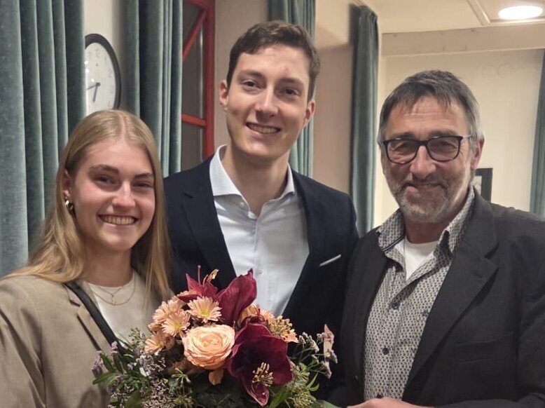 Drei Personen lächeln, eine hält einen Blumenstrauß. Im Hintergrund Vorhänge.