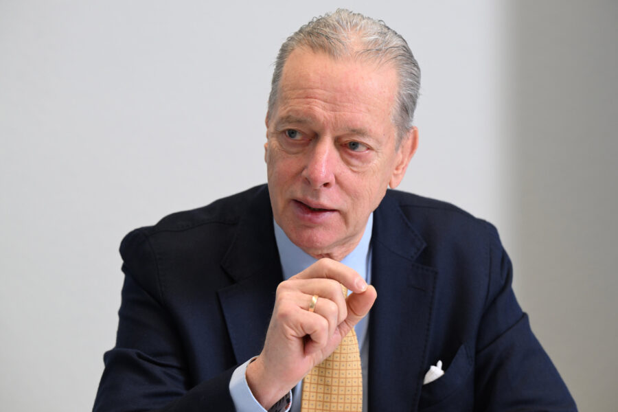 Älterer Mann im Anzug gestikuliert mit der Hand, neutraler Hintergrund.