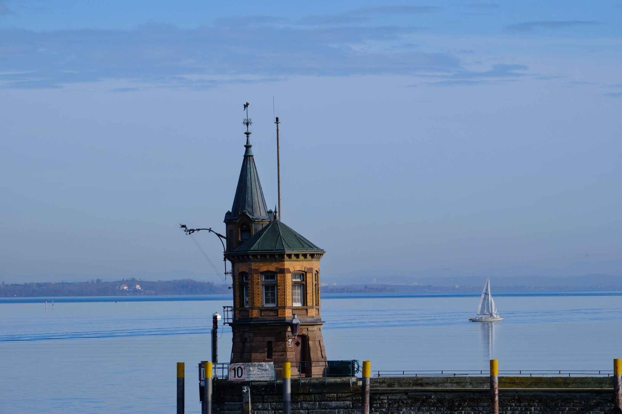 Turm am Wasser, Segelboot im Hintergrund, blauer Himmel.