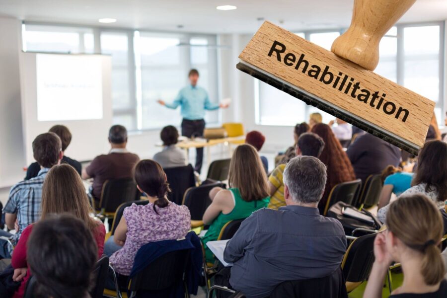 Personen in einem Seminarraum, großer Stempel mit "Rehabilitation".