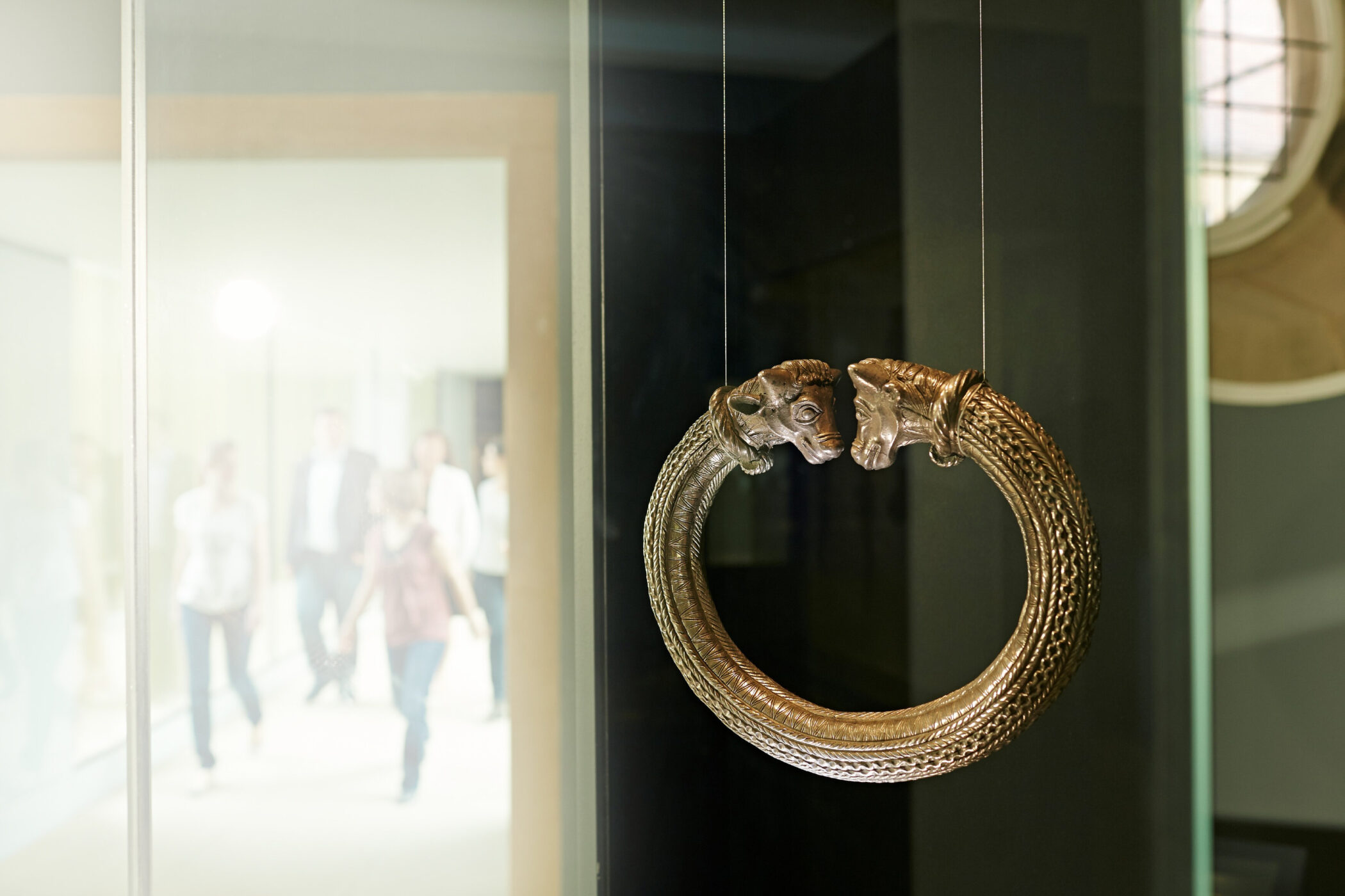 Runder, verzierter Armreif mit Tierköpfen, im Hintergrund unscharfe Personen.