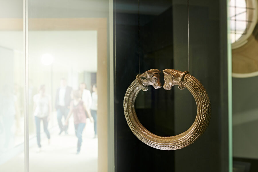 Runder, verzierter Armreif mit Tierköpfen, im Hintergrund unscharfe Personen.