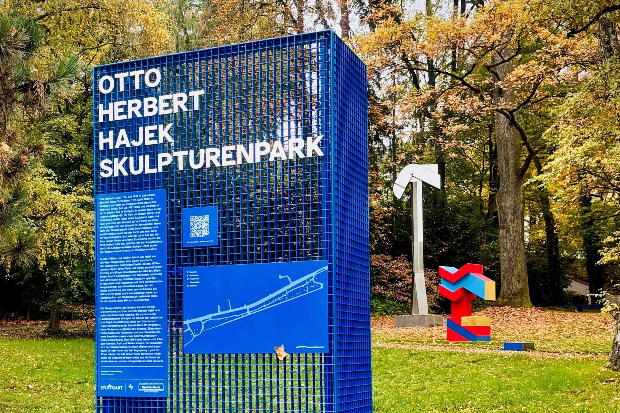 Blaues Schild mit "Otto Herbert Hajek Skulpturenpark", Bäume und Skulpturen im Hintergrund.