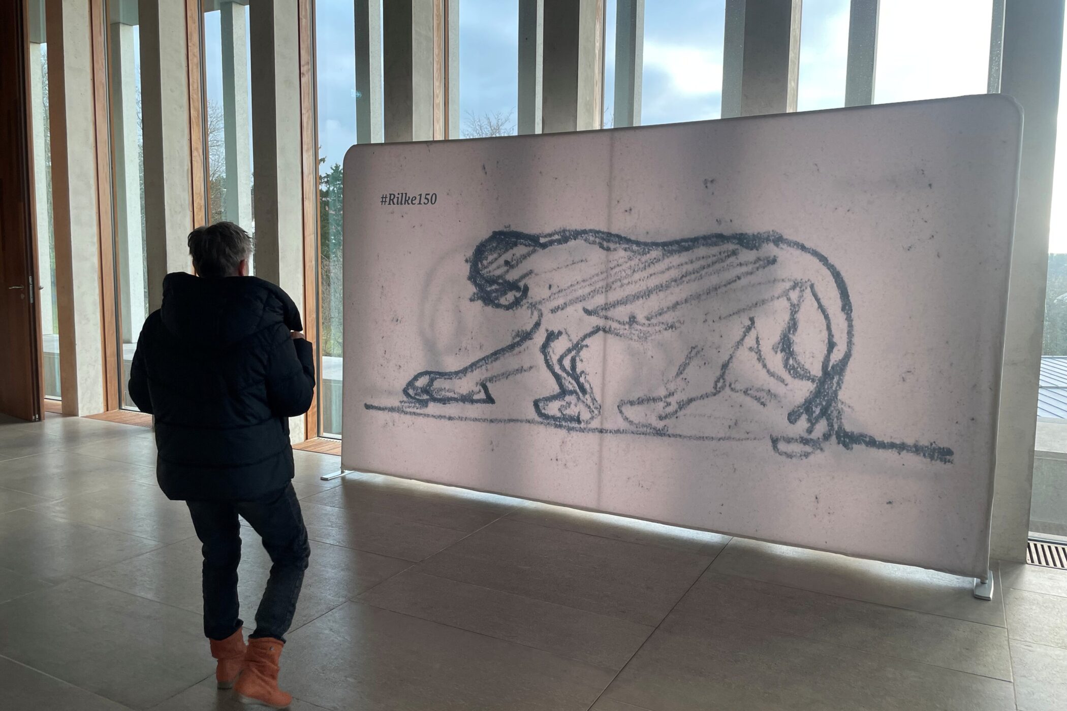 Person betrachtet Zeichnung eines Panthers mit #Rilke150 in einem Raum mit Fenstern.