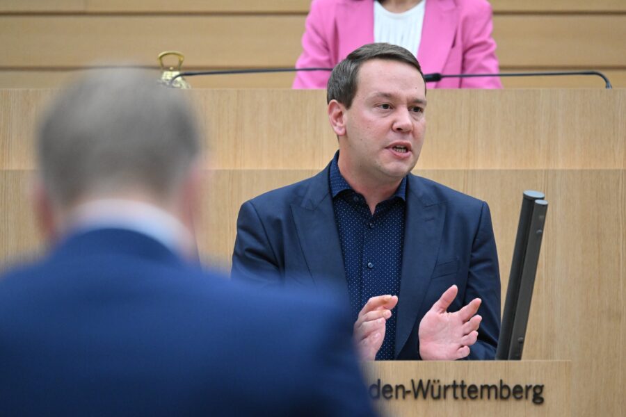 Mann spricht in einem Saal, im Hintergrund eine Person in pinkem Blazer.