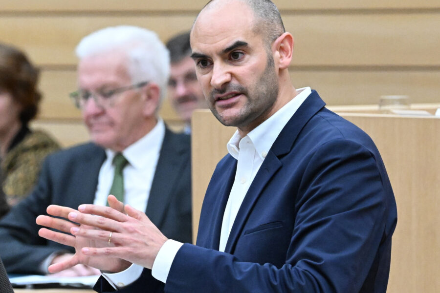 Ein Mann im Anzug spricht gestikulierend in einem Saal. Weitere Personen im Hintergrund.