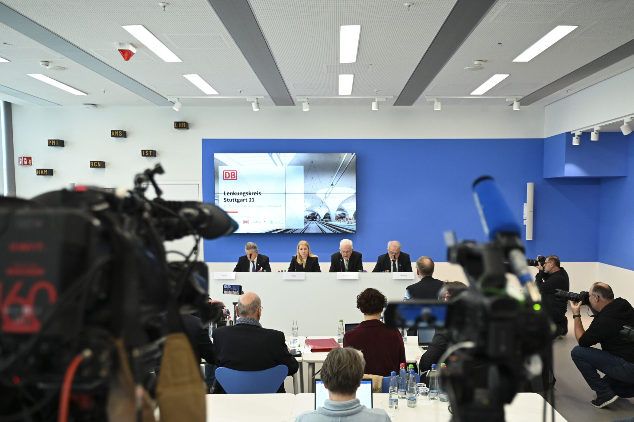 Vier Personen sitzen bei einer Pressekonferenz vor einem Bildschirm.