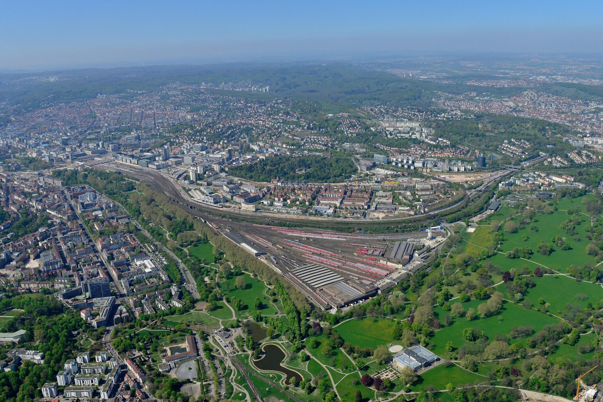 Luftaufnahme einer Stadt mit Bahngleisen, Gebäuden und großen Grünflächen.