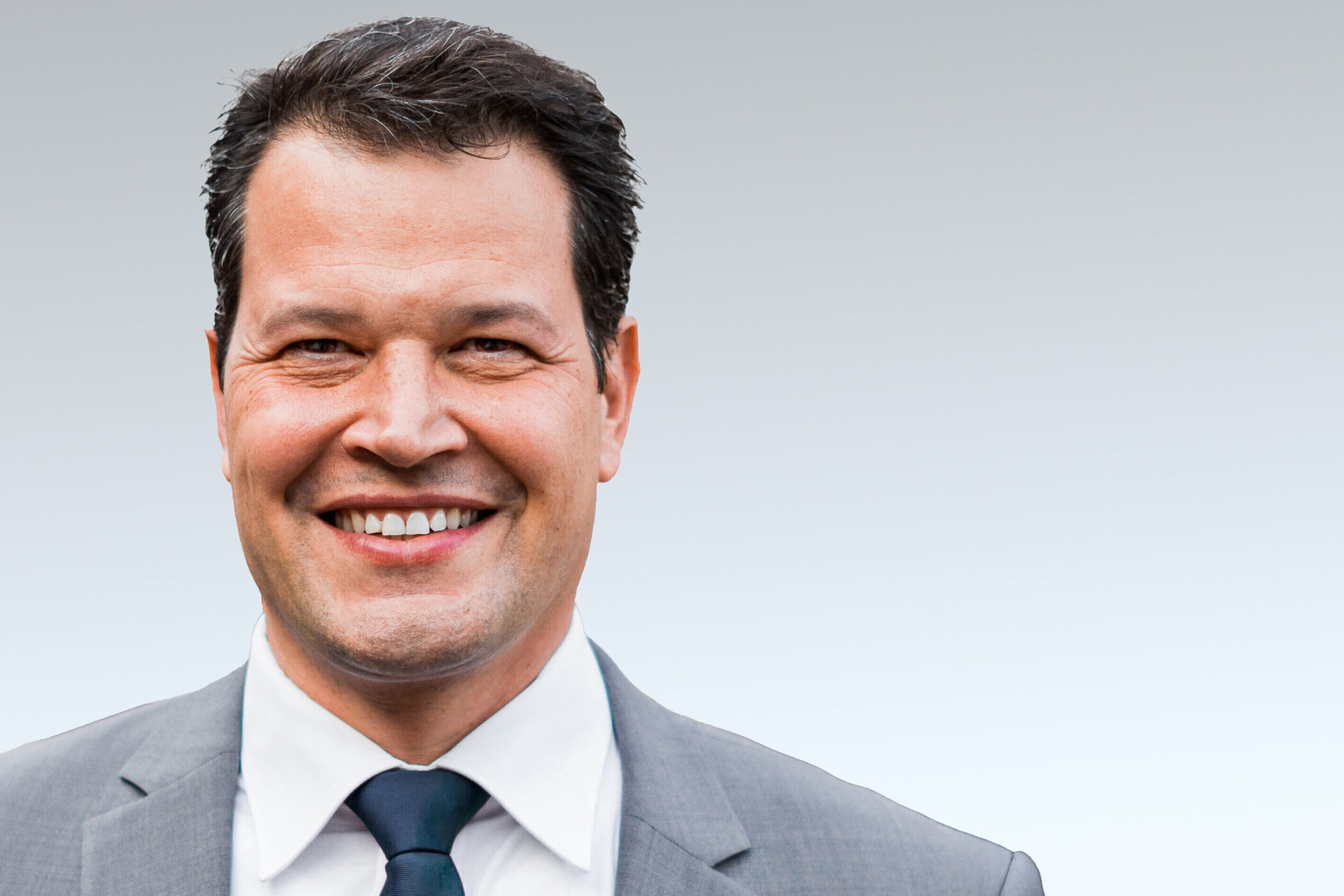 Lächelnder Mann in Anzug mit Krawatte vor grauem Hintergrund.