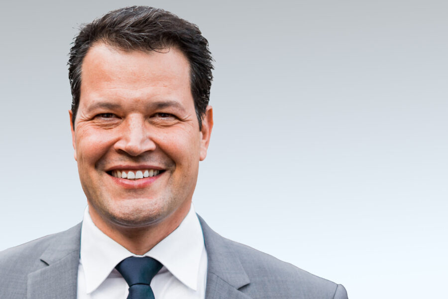 Lächelnder Mann in Anzug mit Krawatte vor grauem Hintergrund.