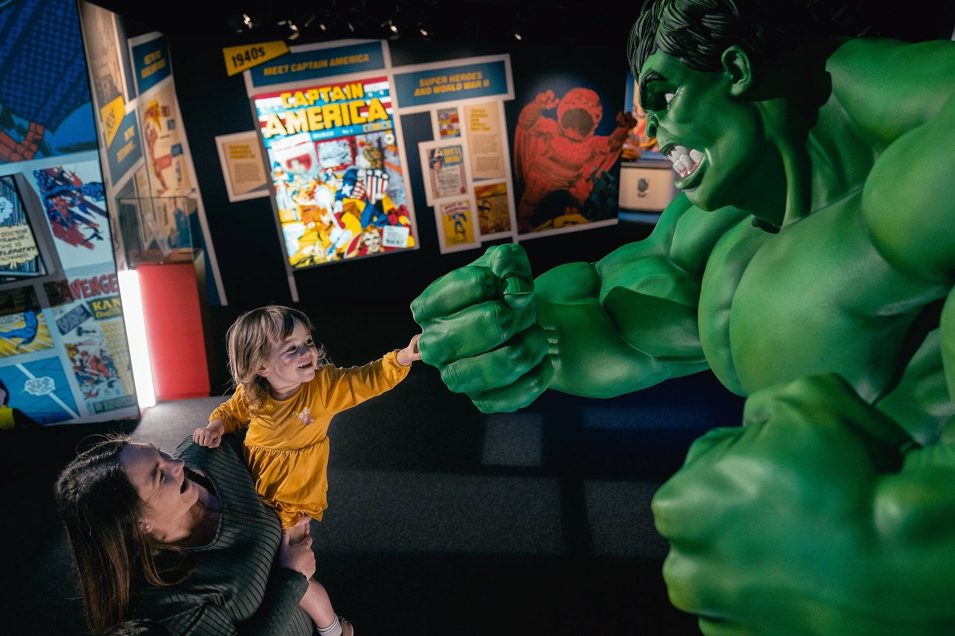 Kind in gelbem Kleid berührt Hulk-Statue, Frau hält das Kind. Comic-Poster im Hintergrund.