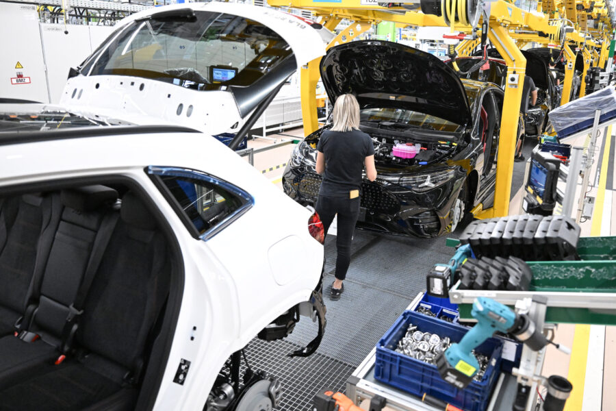 Frau arbeitet an schwarzem Auto in Fabrikhalle, weiße Karosserie im Vordergrund.