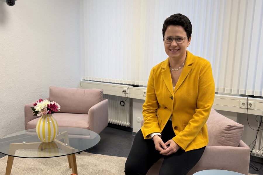 Person in gelbem Blazer sitzt auf rosa Sessel, daneben Glastisch mit Vase und Blumen.