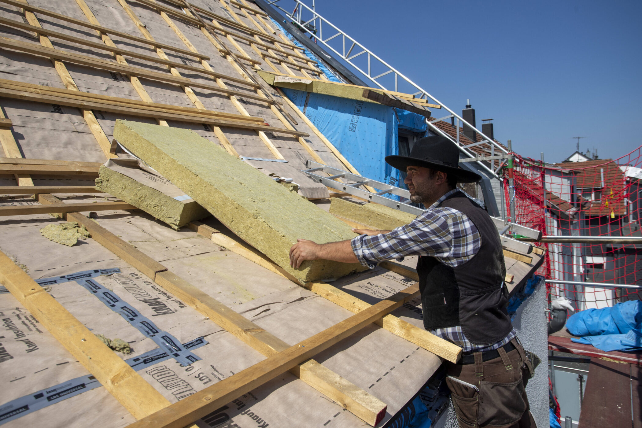 Person legt Dämmplatten auf ein Dach, trägt Hut und Arbeitskleidung.