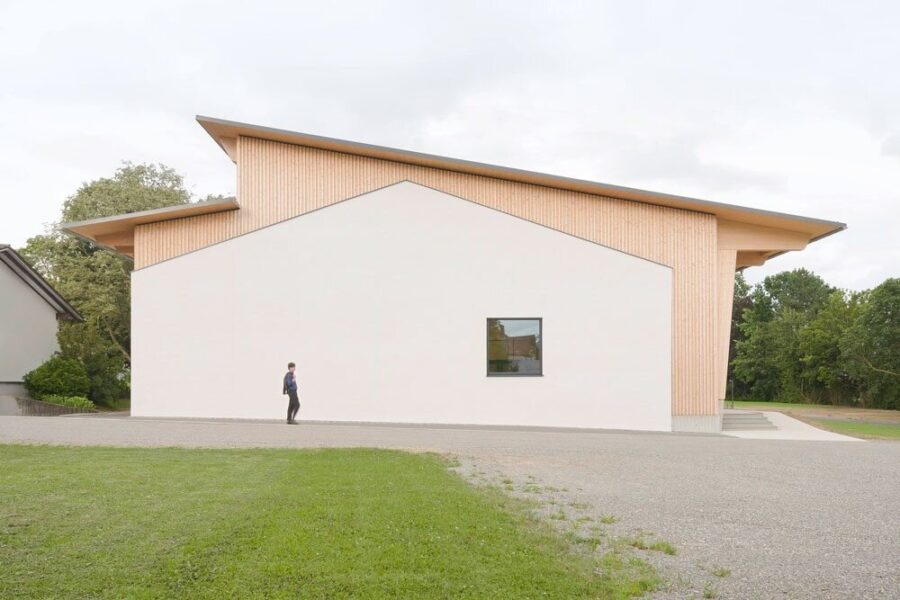 Person geht an modernem Gebäude mit schrägem Dach und kleinem Fenster vorbei.