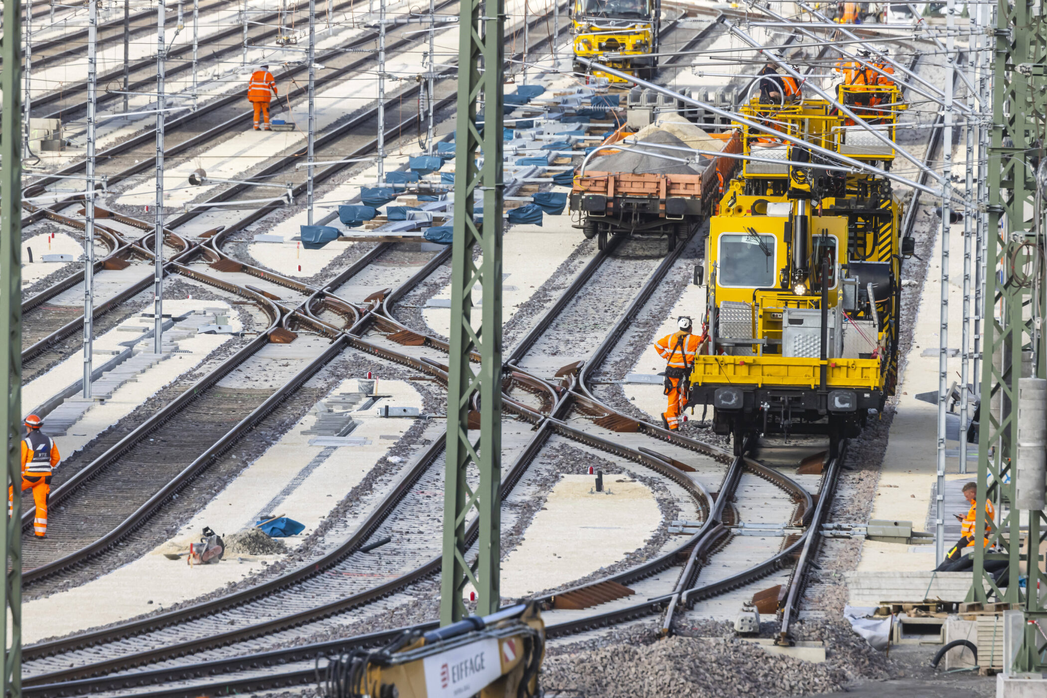 Mehrere Arbeiter in orangefarbener Kleidung auf Bahngleisen mit gelben Maschinen.