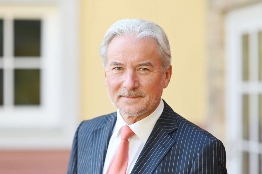 Älterer Mann im Anzug mit Krawatte, lächelt leicht, unscharfer Hintergrund.