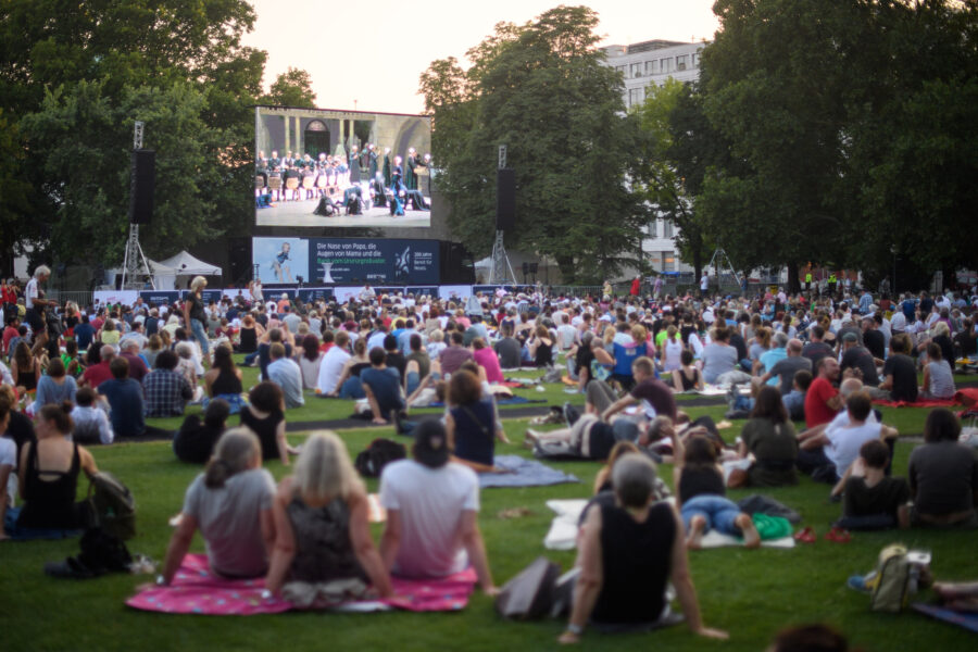 Viele Menschen sitzen auf einer Wiese und schauen auf eine große Leinwand.