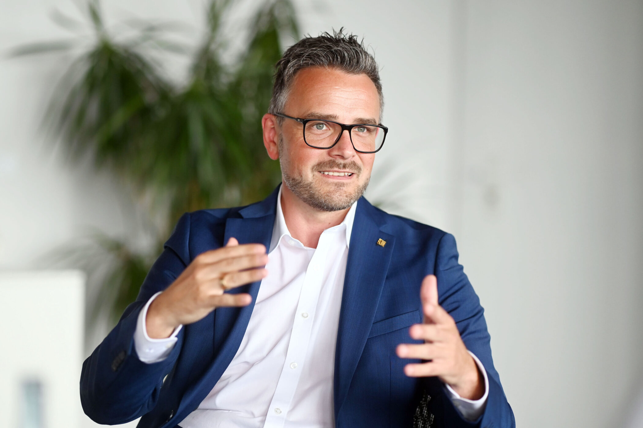 Mann mit Brille und Bart in blauem Anzug gestikuliert, im Hintergrund Pflanzen.