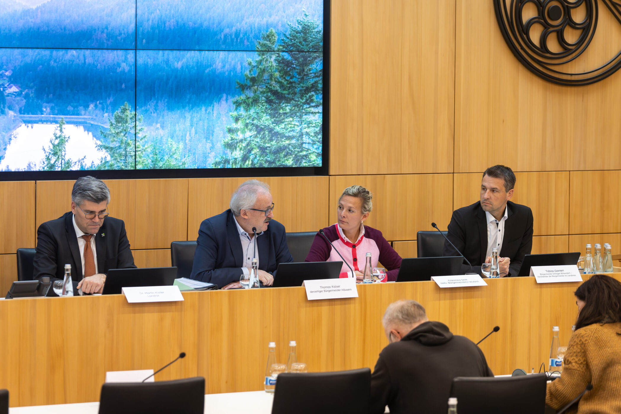 Vier Personen sitzen an einem Tisch mit Laptops, im Hintergrund ein Waldmotiv.