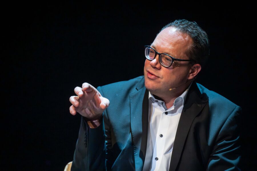 Mann in Anzug mit Brille spricht, Hand erhoben, dunkler Hintergrund.