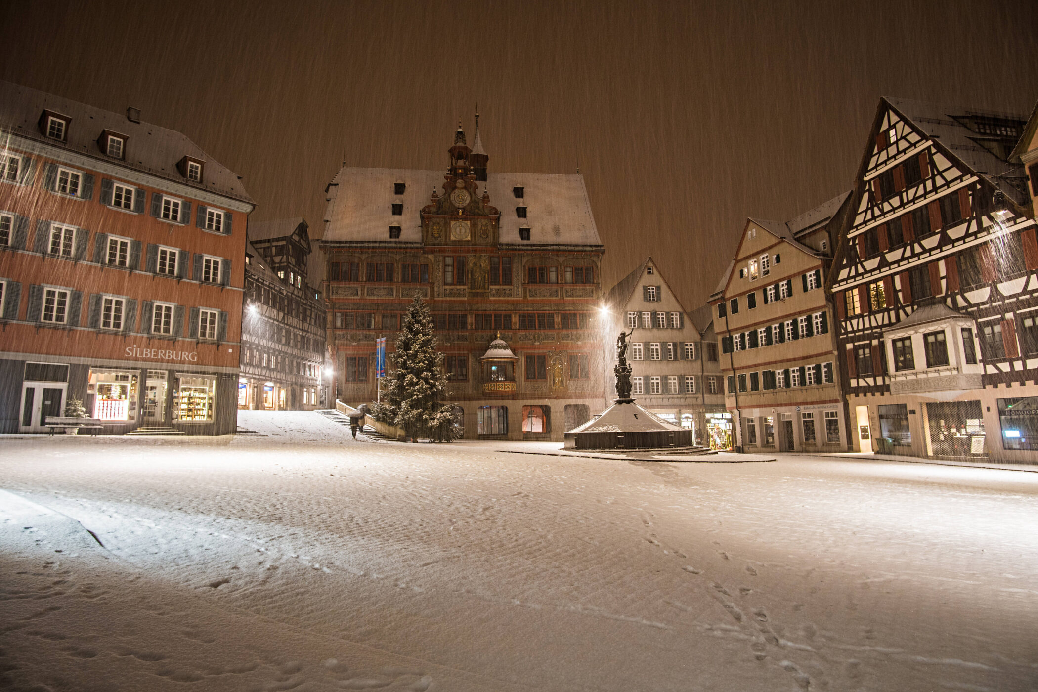 Verschneiter Platz mit Fachwerkhäusern und beleuchtetem Brunnen bei Nacht.