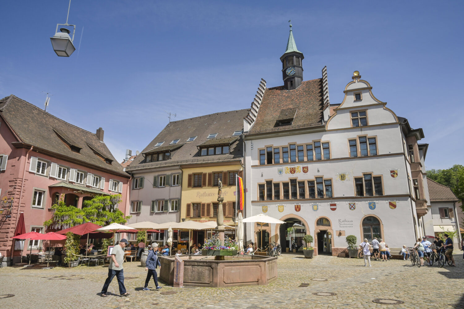 Historische Gebäude mit Uhrturm, Menschen spazieren, Sonnenschirme und Brunnen auf dem Platz.