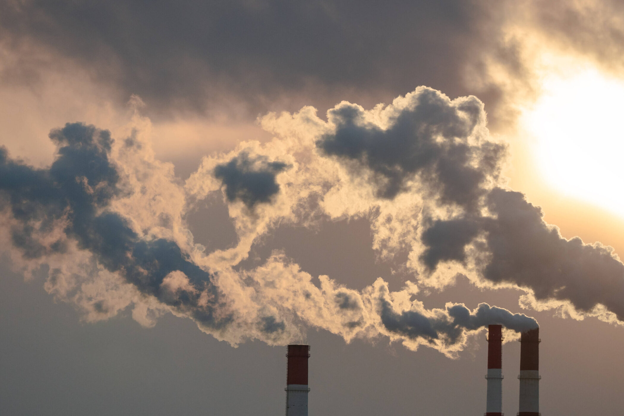 Drei Schornsteine stoßen Rauch in den Himmel vor untergehender Sonne aus.