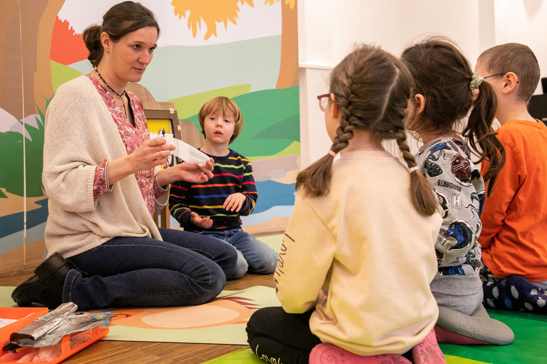 Fünf Personen sitzen auf dem Boden, eine Frau zeigt etwas. Kinder schauen zu.