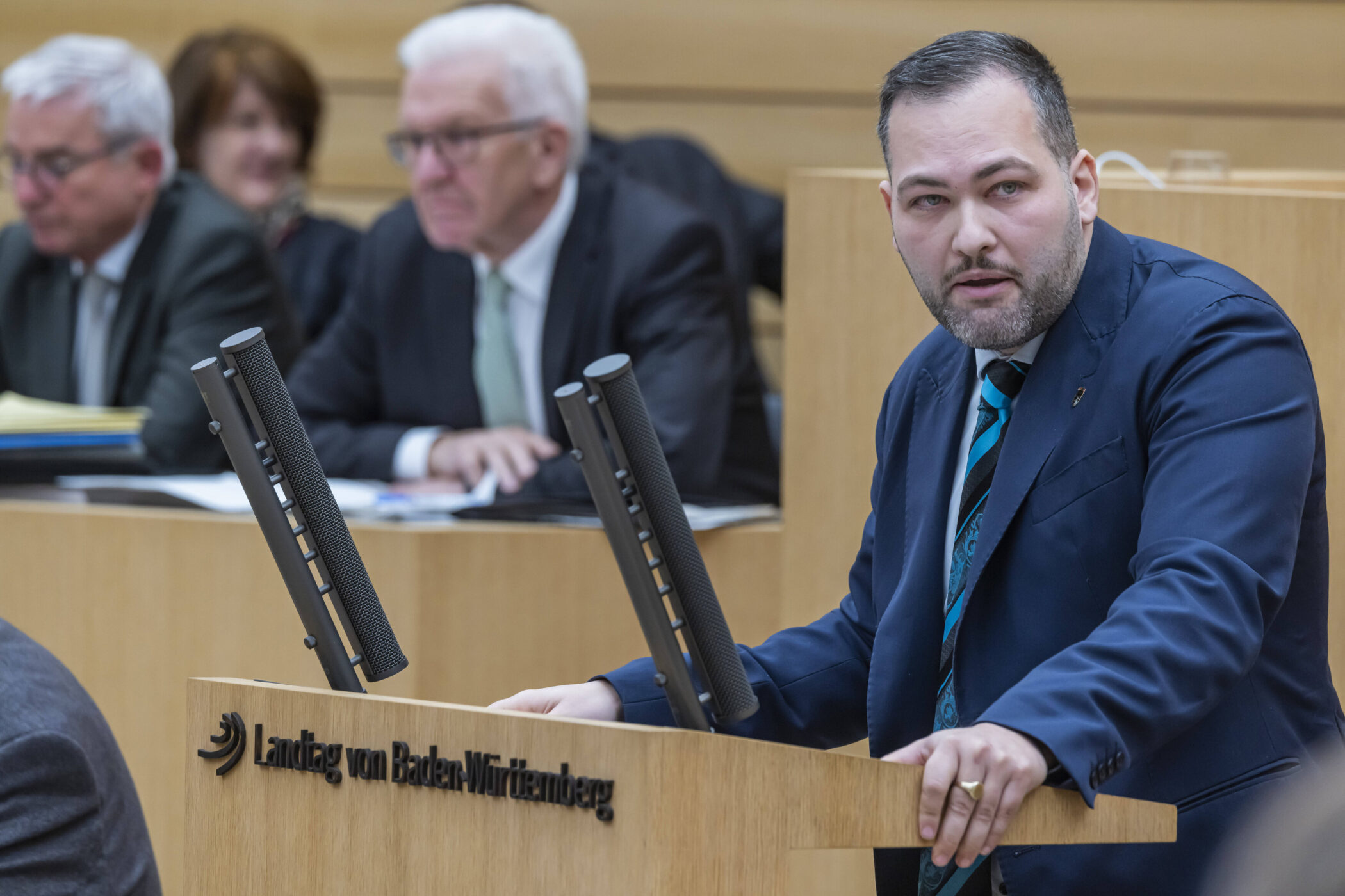 Mann spricht am Rednerpult im Landtag von Baden-Württemberg, mehrere Personen im Hintergrund.