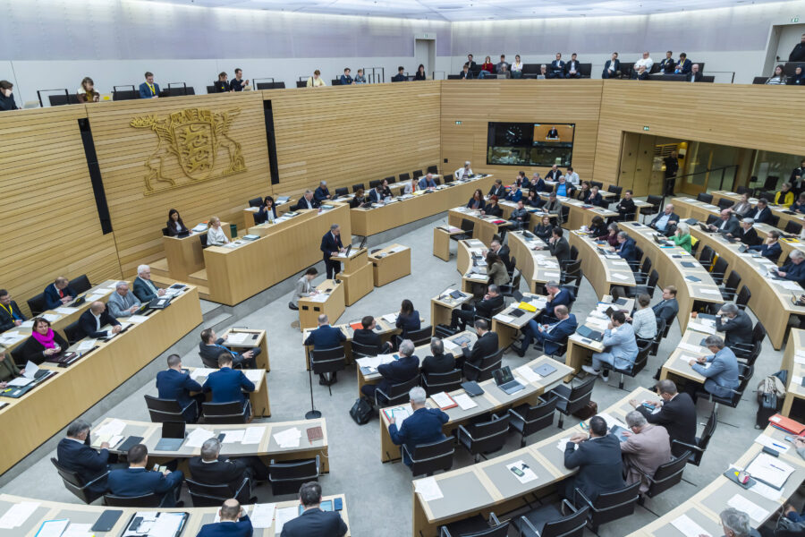 Versammlung in einem großen, modernen Sitzungssaal mit vielen Menschen an Tischen.