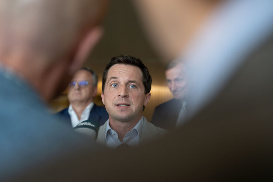 Drei Männer in Anzügen, einer spricht in Mikrofone, unscharfe Köpfe im Vordergrund.