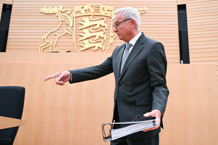 Mann im Anzug zeigt auf Stuhl, hält Aktenordner, Wappen im Hintergrund.