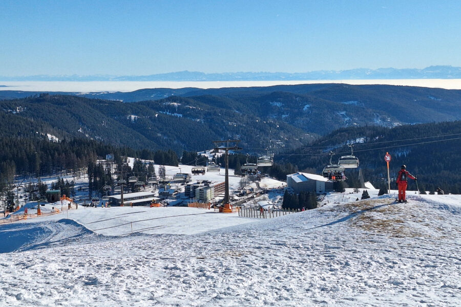 Verschneite Berglandschaft mit Skilift und Skifahrern, bewaldete Hügel im Hintergrund.