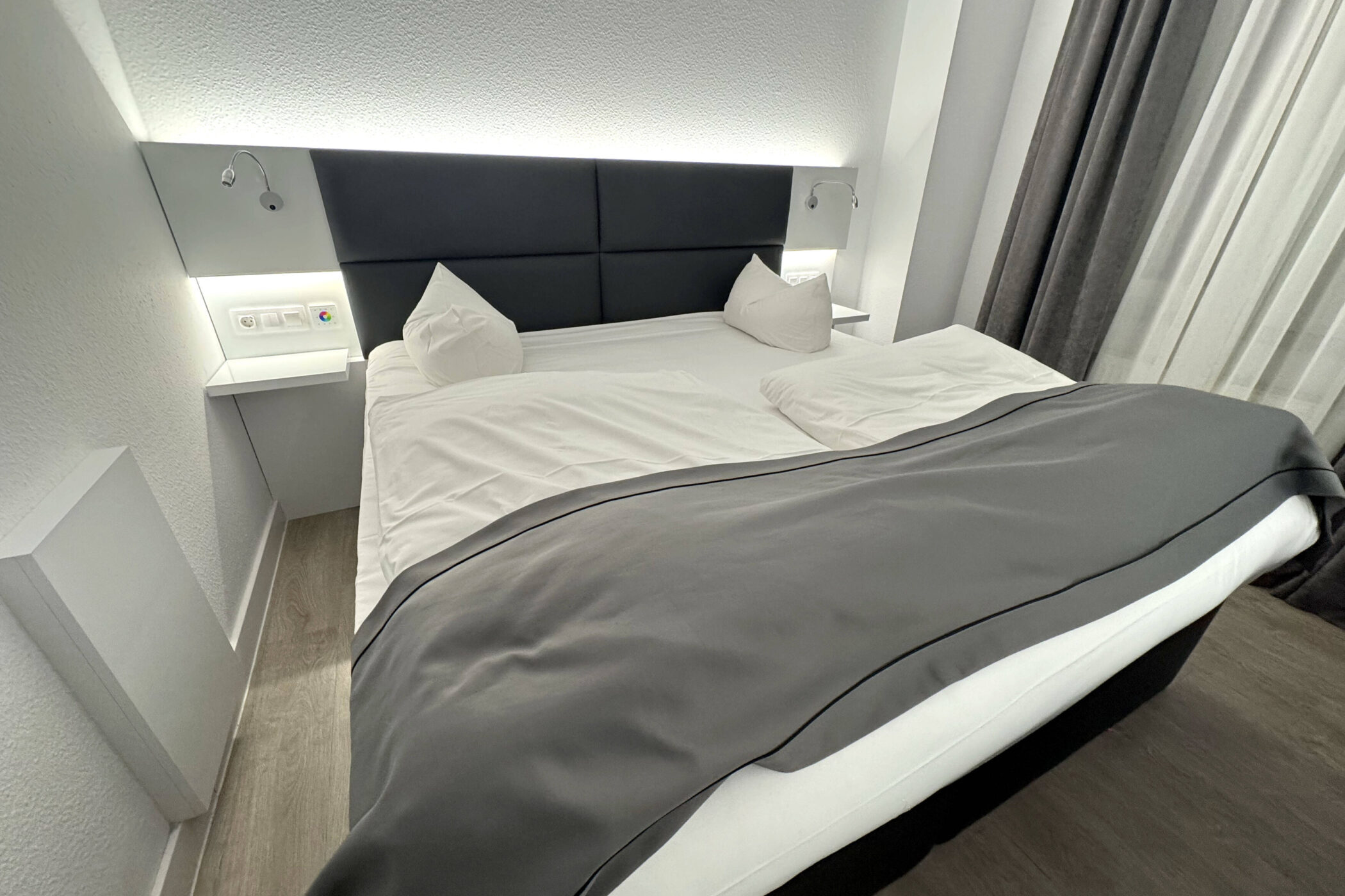 Doppelbett mit weißer Bettwäsche, grauer Decke, zwei Kissen, Wandlampen und Vorhängen.