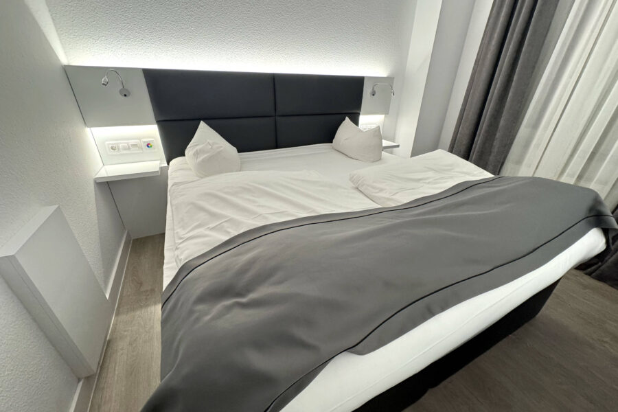 Doppelbett mit weißer Bettwäsche, grauer Decke, zwei Kissen, Wandlampen und Vorhängen.