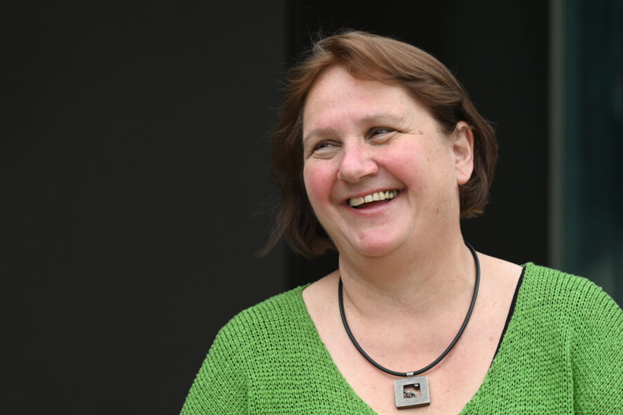 Lachende Frau mit grünem Oberteil und Halskette vor dunklem Hintergrund.