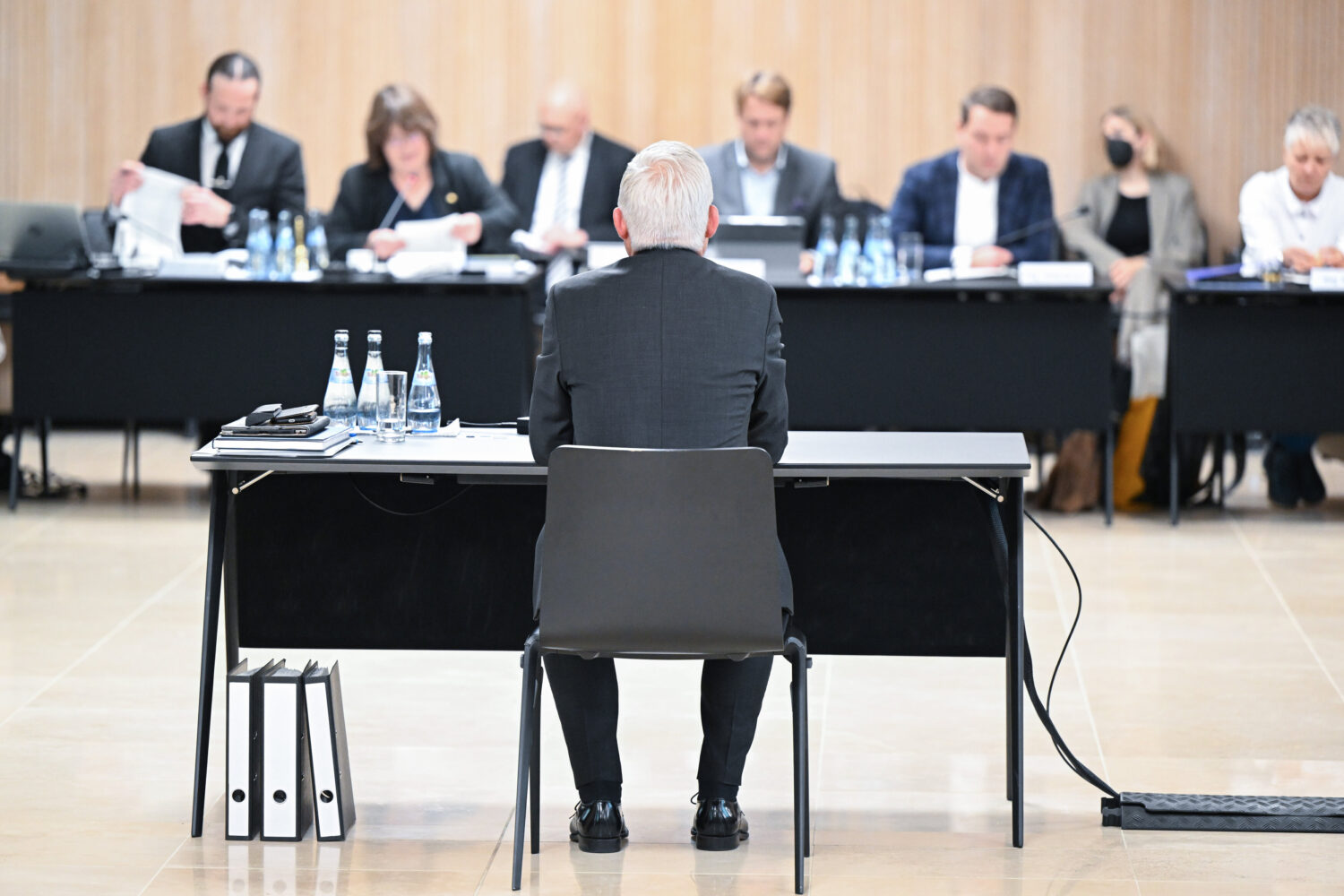 Mann sitzt an Tisch, vor ihm Aktenordner; mehrere Personen gegenüber an Tischen.