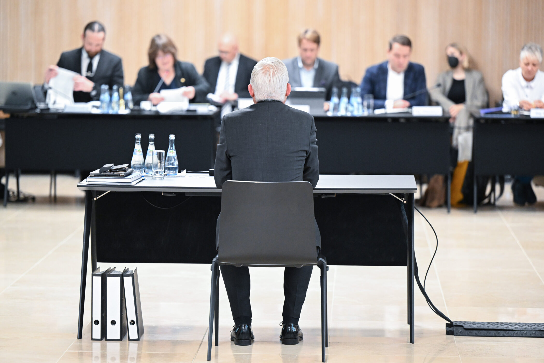 Mann sitzt an Tisch, vor ihm Aktenordner; mehrere Personen gegenüber an Tischen.