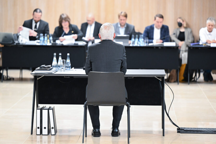 Mann sitzt an Tisch, vor ihm Aktenordner; mehrere Personen gegenüber an Tischen.