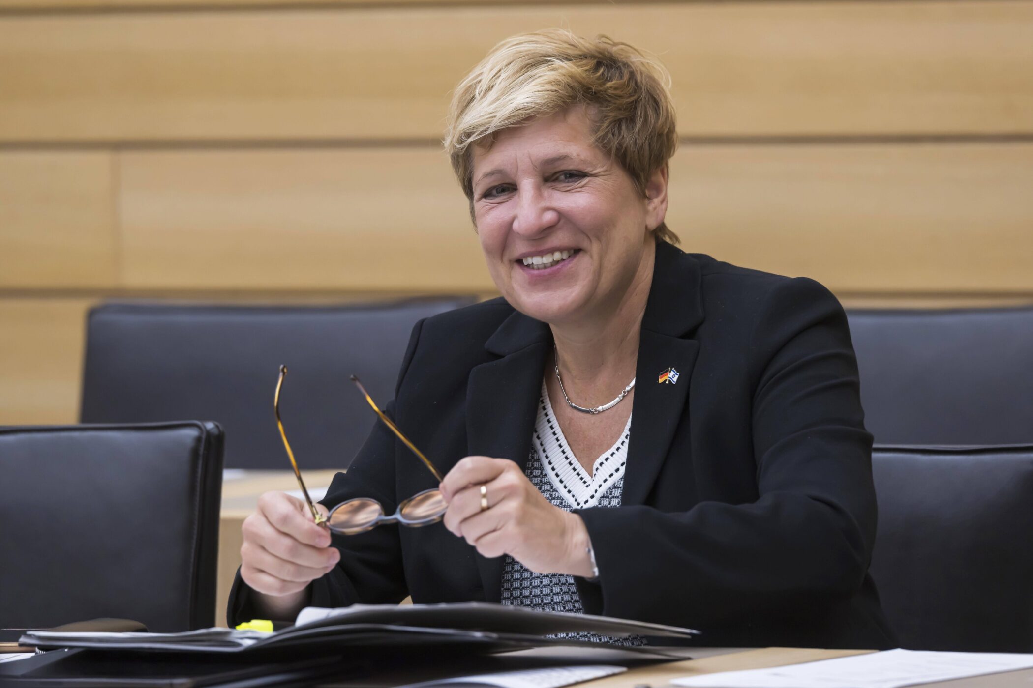 Lächelnde Person im Anzug hält eine Brille, sitzt an einem Tisch mit Papieren.