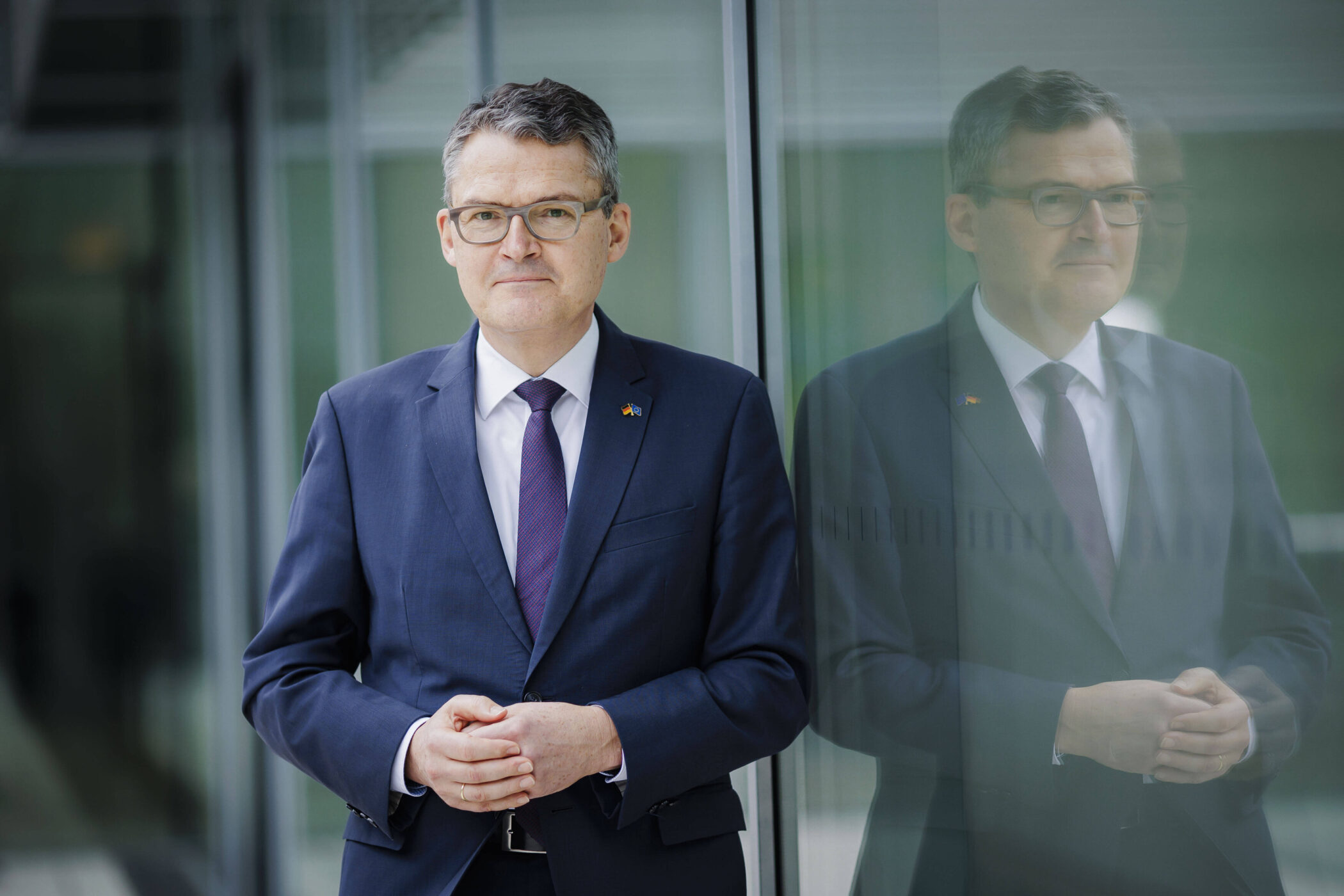 Mann im Anzug steht vor Glaswand, Spiegelbild sichtbar.