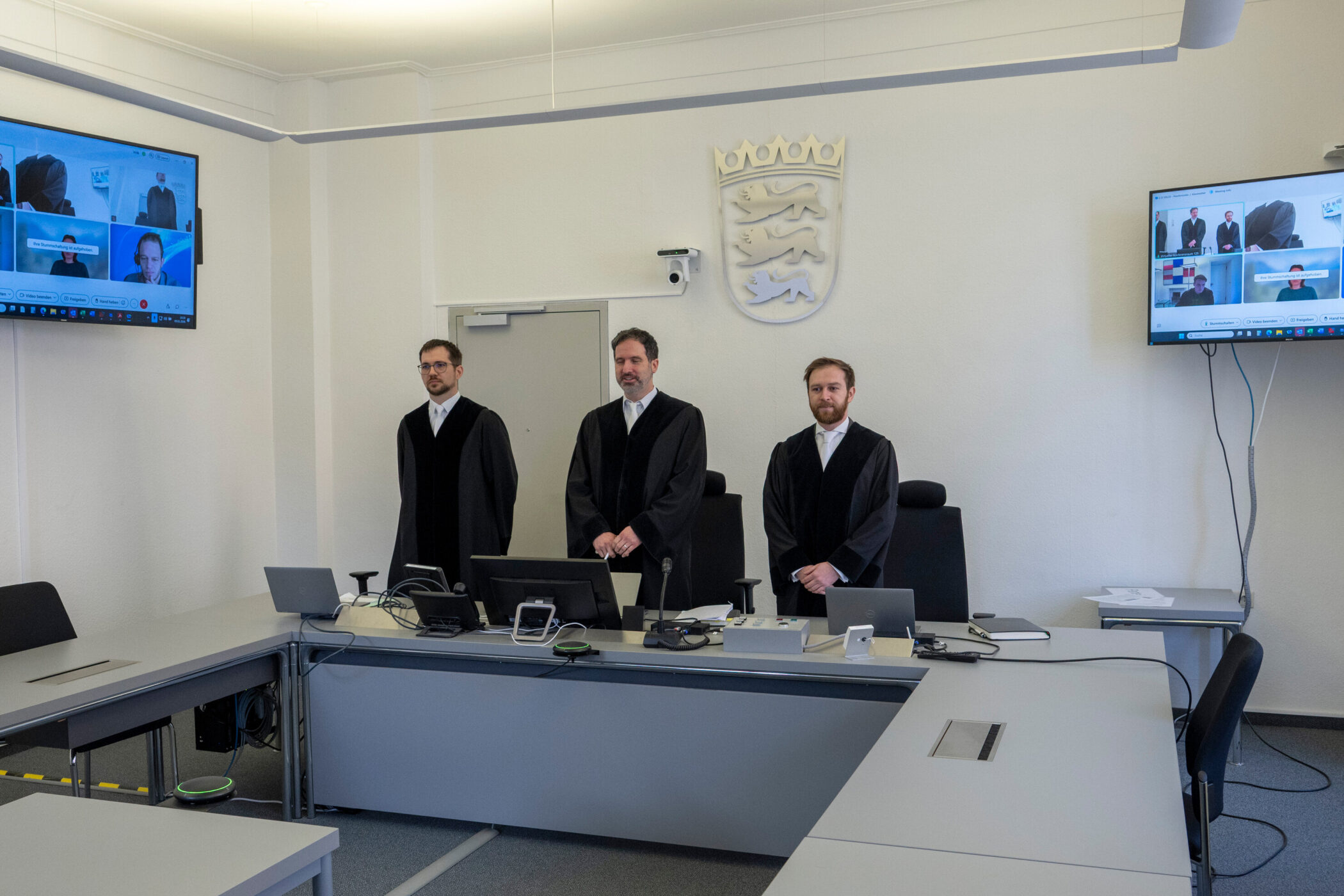 Drei Männer in Roben stehen in einem Raum mit zwei Bildschirmen und einem Wappen an der Wand.