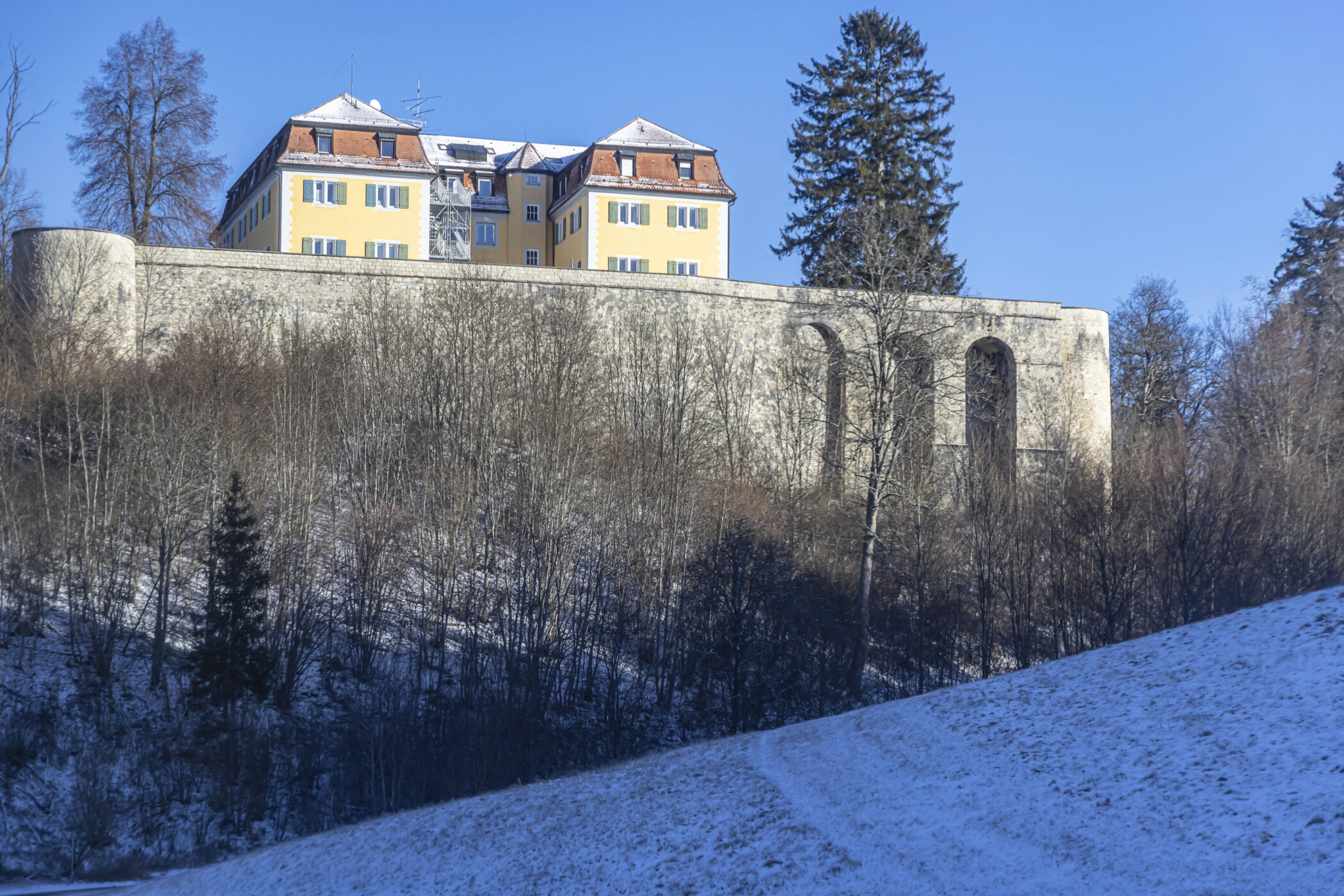 Gelbes Gebäude mit roten Dächern auf einer Mauer, umgeben von schneebedeckten Bäumen.