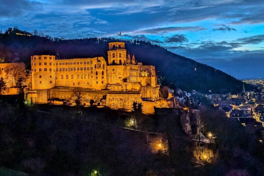 Beleuchtetes Schloss auf Hügel bei Nacht, Stadt im Hintergrund, blauer Himmel.