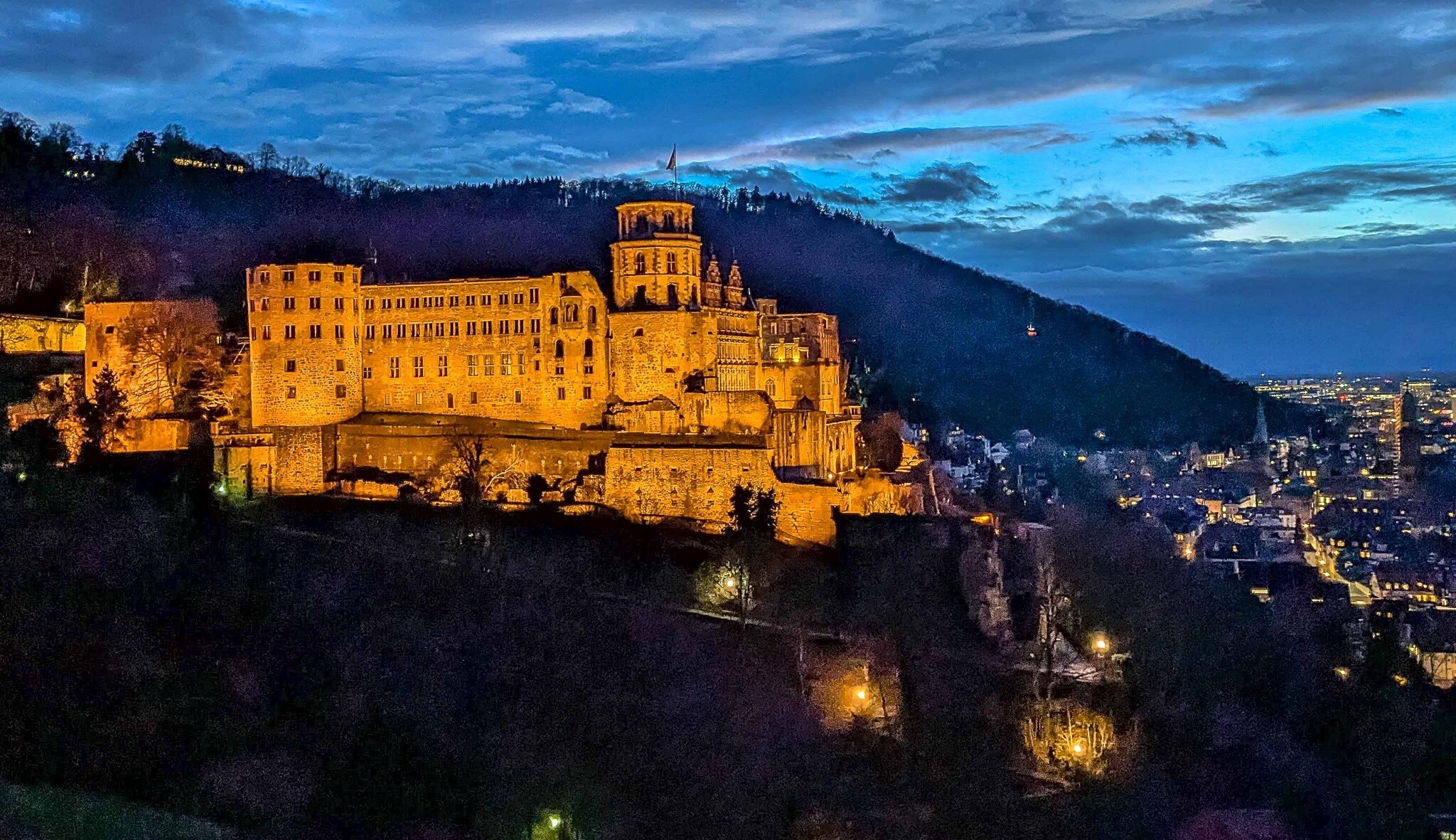 Beleuchtetes Schloss auf Hügel bei Nacht, Stadt im Hintergrund, blauer Himmel.