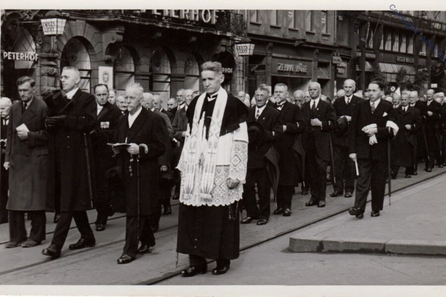 Gruppe von Männern in Anzügen und ein Priester in einer Prozession auf der Straße.