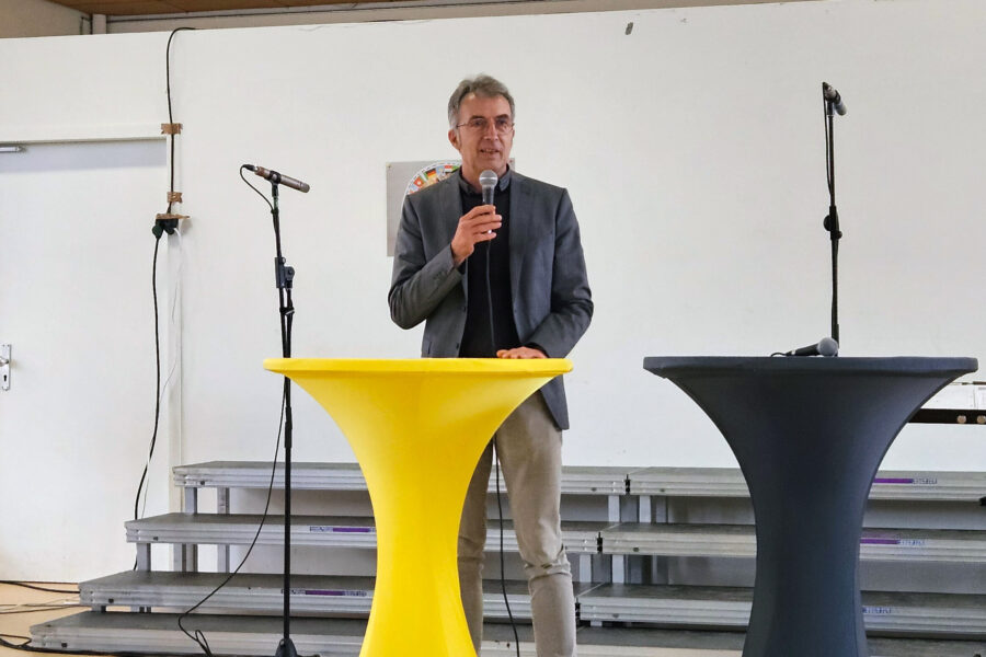 Mann spricht in Mikrofon, steht zwischen gelbem und schwarzem Stehtisch.